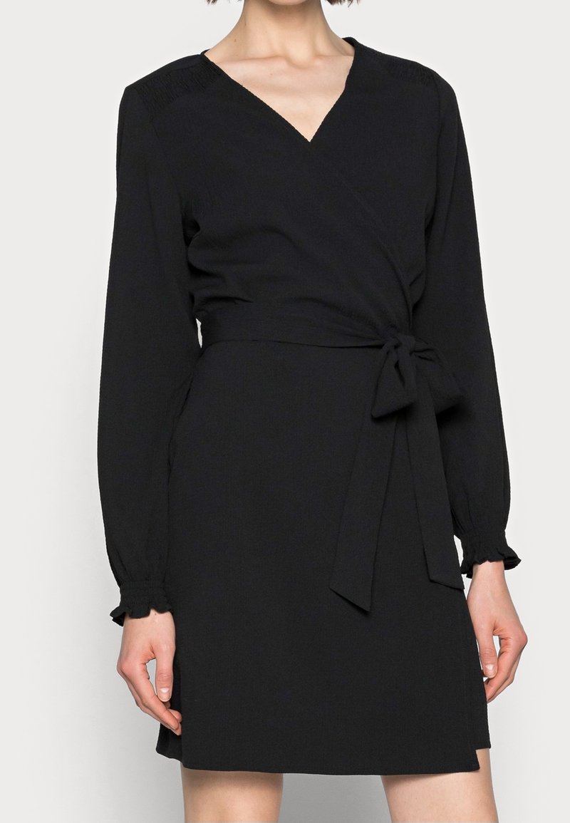 Femme portant une robe portefeuille noire à manches longues avec un décolleté en V et une ceinture nouée à la taille.
