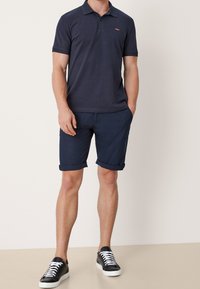 Granatowa koszulka polo z kołnierzykiem, zestawiona z granatowymi szortami z podwiniętymi nogawkami. Czarne sneakersy z białymi sznurowadłami. Prosty, casualowy design i krój.