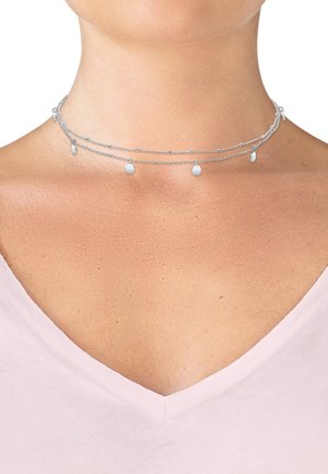 Silver multi-layered choker necklace featuring delicate chains, round pendants, and small beads. Worn against a light pink top.