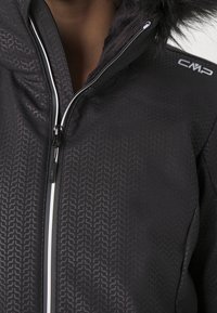 Gros plan d'une veste noire à motifs avec fermeture éclair blanche, col doublé en fourrure et logo CMP sur l'épaule.