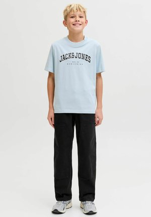 Garçon blond debout et souriant, portant un T-shirt bleu clair "JACK&JONES", un pantalon noir à poches avant et des baskets grises.