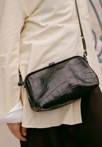 Borsa a tracolla nera in pelle sintetica effetto coccodrillo, di forma rettangolare, con chiusura superiore e tracolla regolabile, indossata sopra una camicia a righe gialle.