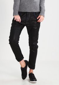 Černé kalhoty s koženým vzhledem a zúženým střihem, s předními kapsami a dekorativním šitím. Kombinované s černými teniskami a šedým pleteným svetrem.