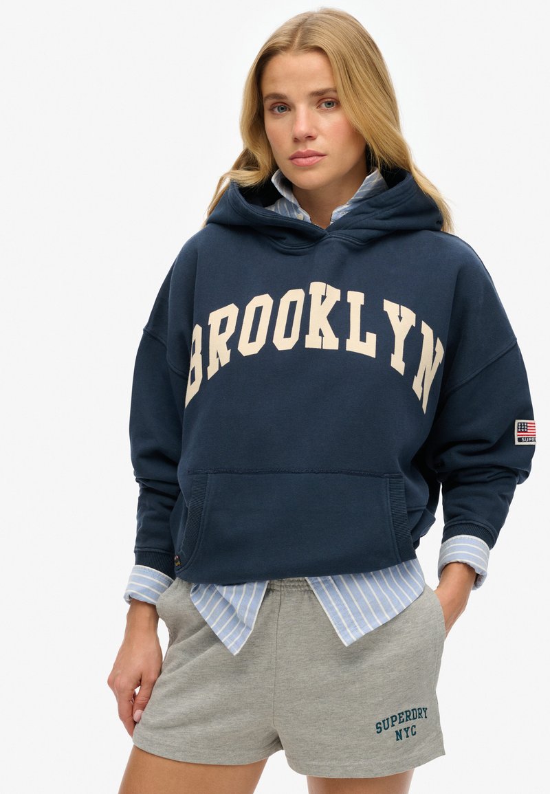 Marine sweatshirt met capuchon en "BROOKLYN" in crèmekleurige letters, voorzak en geribbelde boorden. Grijze sportshorts met "SUPERDRY NYC" in groene borduurwerk.