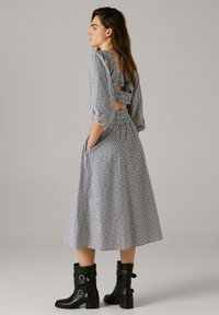 Žena u midi haljini s prugama, napuhanih rukava i otvorenih leđa, rukama u džepovima, nosi crne gležnjače s kopčom, stoji bočno.