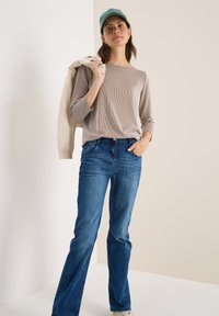 Beige ribbad långärmad topp i kombination med utsvängda blåjeans. En ljusbeige jacka hänger över axeln, kompletterad med en turkos keps.