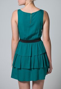 Türkisfarbenes Kleid mit ärmellosem Design, geschichteten Rüschen am Rock und einem goldenen Reißverschlussdetail auf der Rückseite. Schwarzer Taillenbund als Akzent.