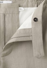 Pantalon beige clair avec fermeture éclair, boutonnière et doublure intérieure blanche, mettant l'accent sur la ceinture et les détails de la fermeture.