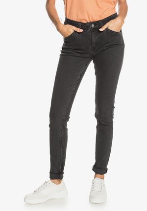 Kapeat mustat denim-farkut korkealla vyötäröllä, viiden taskun muotoilu ja käännetyt haitarit, yhdistettyinä valkoisiin lenkkareihin ja oranssiin puseroon.