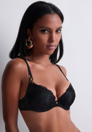 Femme à la peau mate porte un soutien-gorge en dentelle noire et de grandes boucles d'oreilles créoles dorées, posant devant un fond gris clair uni.