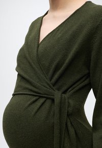 Personne enceinte portant une robe portefeuille texturée vert foncé avec un col en V et une taille nouée, vue de la poitrine jusqu'au ventre.
