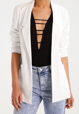 Vrouw draagt een witte blazer over een zwart topje met een diepe V-hals en horizontale bandjes, gecombineerd met lichtblauwe high-waisted jeans.