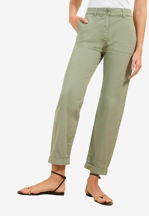 Žena oblečená v svetlo olivových rovno strihaných nohaviciach s vyhrnutými lemami a čiernych plochých sandáloch so zlatým krúžkovým detailom, stojaca s jednou rukou v vrecku.