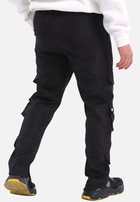 Schwarze Cargo-Hosen mit elastischem Bund, mehreren Seitentaschen und glatter Textur. Kombiniert mit schwarzen Sportschuhen mit gelben Akzenten.