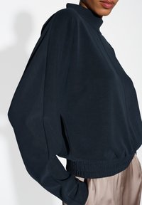 Top corto blu scuro con colletto alto, maniche ampie e cintura elastica, realizzato in un tessuto liscio e leggero.