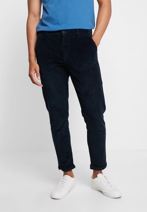 Mann trägt hochgekrempelte schwarze Cordhose, blaues T-Shirt und weiße Sneakers, steht mit einer Hand in der Tasche vor einfarbigem Hintergrund.