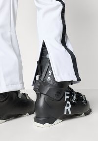 Μαύρα χιονιστικά παπούτσια με ματ φινίρισμα εμφανίζουν ένα ασημί λογότυπο και αριθμούς. Λευκό παντελόνι διαθέτει πλαϊνό φερμουάρ και μαύρες λεπτομέρειες στη ραφή.