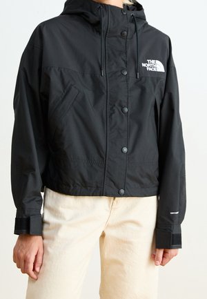 Osoba w czarnej kurtce z kapturem The North Face zapinanej na guziki oraz w jasnobeżowych spodniach na jednolitym tle.