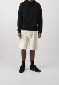 Sweat à capuche noir avec une poche kangourou, associé à un short crème avec poches latérales. Des baskets noires avec des accents gris complètent le look.