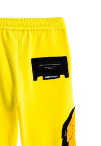 Pantalones de chándal amarillos con un parche de cinta VHS falsa negra en la parte trasera, que presenta texto y un elemento de diseño gráfico a lo largo de la pierna.