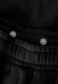 Juodos sportinės kelnės su surinktu elastingu juosmeniu, dviem mygtukais ir tekstūruotu audiniu. Paviršius yra lygus, matomi siuvimo detalės.