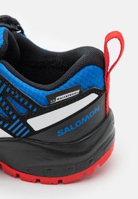 Zapatilla impermeable negra y azul con parte superior de tela texturizada, tirador rojo, suela negra y detalles en blanco con el logo "SALOMON".