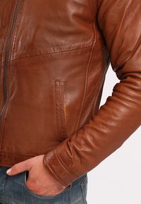 Braune Lederjacke mit Reißverschluss, ausgestattet mit einer Tasche auf der linken Seite und einem taillierten Design, strukturiertem Material und glatter Oberfläche.