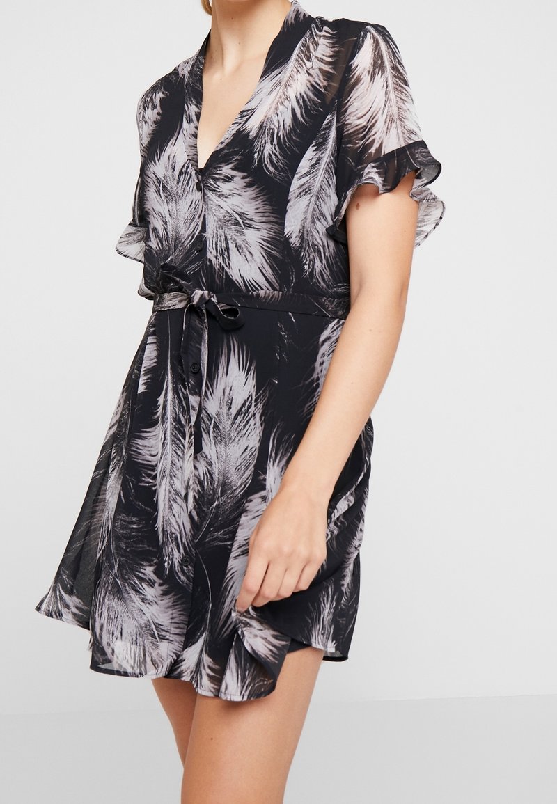 Femme portant une robe courte noire avec des motifs de plumes blanches, des manches à volants, des boutons sur le devant et une ceinture nouée à la taille.