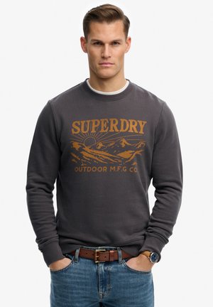 Mand iført mørkegrå sweatshirt med bjerg- og solgrafik samt teksten "SUPERDRY OUTDOOR M.F.G CO", blå jeans, brunt bælte og armbåndsur.