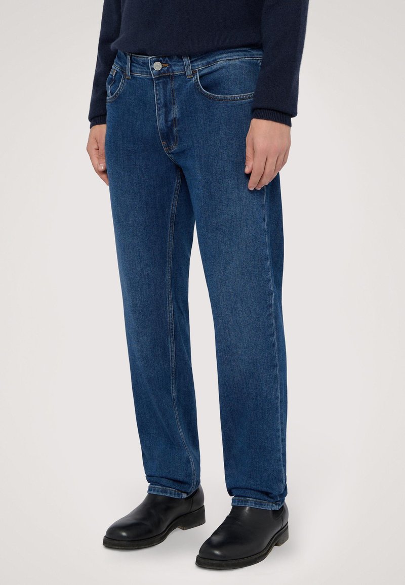 Jeans de denim en azul oscuro con un diseño de pierna recta, cinco bolsillos, costuras sutiles y un cierre frontal de cremallera estándar. Botas negras puestas.