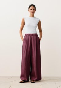 Femeie în picioare cu mâinile în buzunare, purtând un top alb fără mâneci, pantaloni largi burgundy și papuci negri, pe un podea deschisă la culoare.