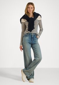 Top a maniche lunghe a righe in crema e blu navy, abbinato a jeans a gamba larga blu chiaro. Maglione blu navy strutturato appoggiato sulle spalle, sneakers bianche.
