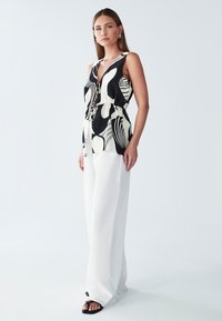 Czarna i biała bezrękawnikowa bluzka w wzory z dekoltem w serek i ściągaczem w pasie, zestawiona z szerokimi białymi spodniami i czarnymi sandałami.