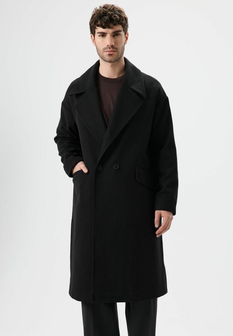 Manteau en laine noire avec col cranté, fermeture à double bouton, et poches latérales. Le mannequin pose devant un fond neutre.