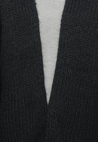 Cardigan nero lavorato a maglia con un profondo scollo a V, caratterizzato da una finitura testurizzata. Indossato sopra una t-shirt grigia chiara.