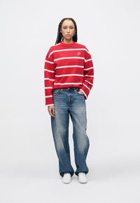 Chemise à manches longues rayée rouge et blanche, coupe décontractée, associée à un jean large bleu clair. Des baskets blanches complètent la tenue.