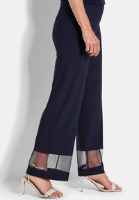 Marineblauwe pantalon met wijde pijpen heeft doorschijnende meshpanelen aan de zoom. Gepaard met zilveren haksandalen, wat het algehele ontwerp versterkt.