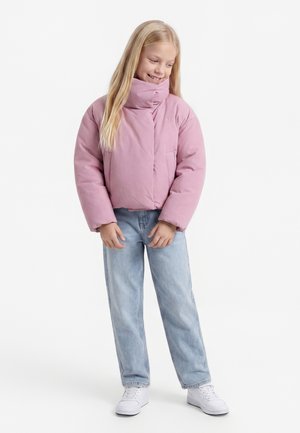 Jeune fille souriante, portant une doudoune rose clair, un jean bleu clair et des baskets blanches, debout devant un fond blanc.