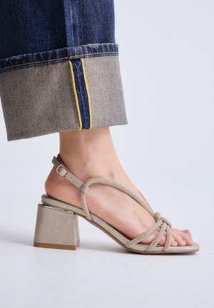 Pie con sandalia de tacón de bloque beige con tiras y hebilla en el tobillo, combinada con vaqueros azules con dobladillo, sobre un fondo claro y liso.