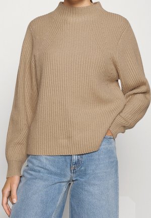 Personne portant un pull en maille côtelée beige à manches longues et un jean bleu clair, une main dans la poche, sur un fond uni.