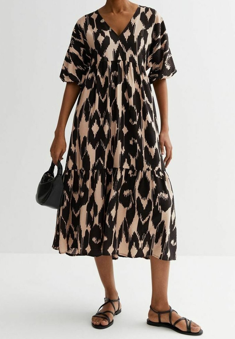 Femme portant une robe midi beige et noire à motif abstrait, des sandales noires à brides, et tenant un petit sac à main noir à ses côtés.