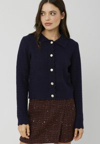Cardigan bleu marine avec un ourlet et un col festonnés, présentant un motif en maille texturée et des fermetures à boutons dorés sur le devant.