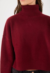 Bordeauxfarbener, gestrickter Rollkragenpullover mit geripptem Kragen und Saum, aus einem weichen, strukturierten Stoff gefertigt. Cropped-Design mit weiten Ärmeln.