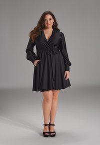 Vestido negro ajustado con mangas largas abullonadas, cintura entallada y falda fluida. Tejido suave con bolsillos, combinado con tacones de plataforma negros.