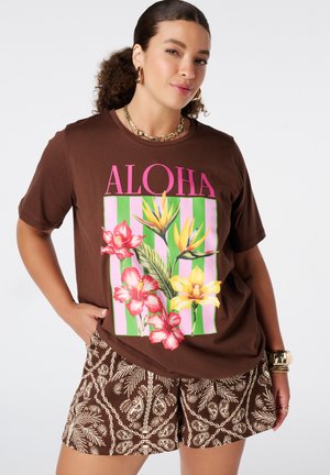 Vrouw draagt een bruine "ALOHA" T-shirt met kleurrijke tropische bloemen en patroonbruine shorts, geaccessoriseerd met gouden sieraden.