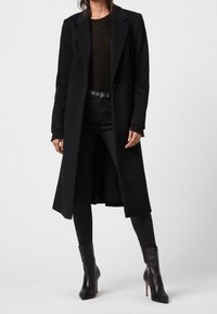 Abrigo largo negro con solapas a juego, cierre de un solo botón, hecho de una mezcla de lana. Se lleva sobre un suéter negro de canalé y jeans ajustados.