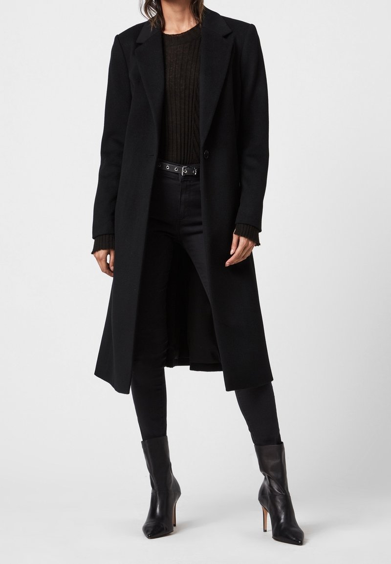 Abrigo largo negro con solapas a juego, cierre de un solo botón, hecho de una mezcla de lana. Se lleva sobre un suéter negro de canalé y jeans ajustados.