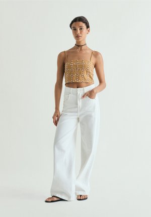 Joven mujer de pie con un top corto marrón con estampado, pantalones blancos de pata ancha, sandalias negras y un collar tipo choker sobre un fondo liso.