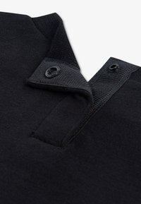 Melns polo apkakle ar gludu faktūru, kas aprīkota ar diviem dekoratīviem melniem aizdares sprādzēm. Audums izskatās mīksts un viegls.