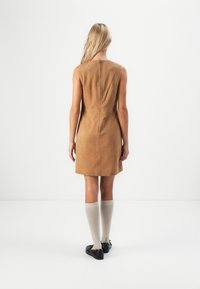 Žena stoji okrenuta leđima, obučena u bezrukavu haljinu od taupe suede, s bež čarapama do koljena i crnim cipelama, naspram obične bijele pozadine.